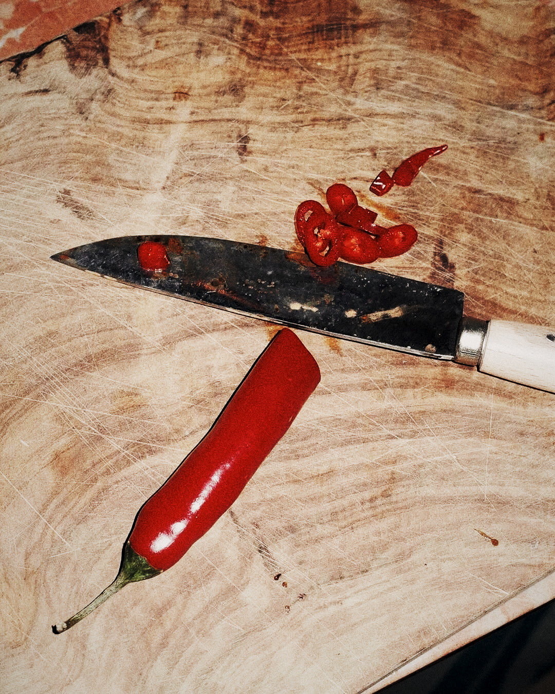 A red chilli and knife on a scarred chopping board — muted colour film, the only saturated colour is red